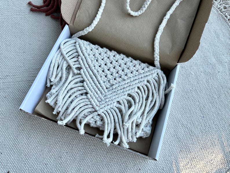 May include: An off-white macrame crossbody bag featuring a triangular flap, intricate knotted cord patterns, and long fringe detailing along the bottom edges. The bag includes a braided shoulder strap and is presented inside an open cardboard box.