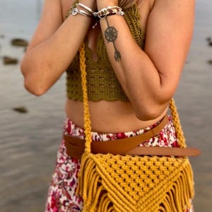 May include: A mustard yellow macrame crossbody bag featuring a triangular flap with a knotted diamond pattern and long fringe trim. The bag is suspended by a thick braided cord strap and is worn over a brown leather belt and a floral patterned skirt.