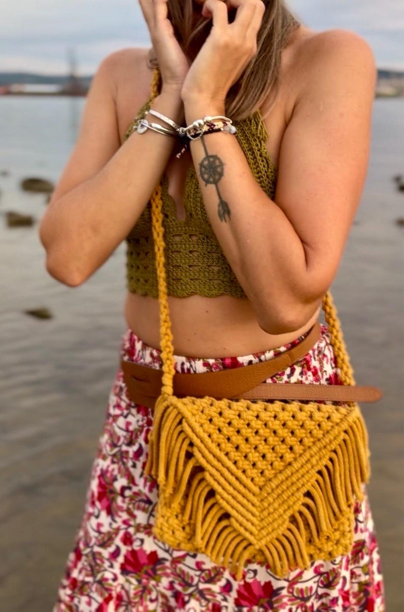 May include: A mustard yellow macrame crossbody bag featuring a triangular flap with a knotted diamond pattern and long fringe trim. The bag is suspended by a thick braided cord strap and is worn over a brown leather belt and a floral patterned skirt.