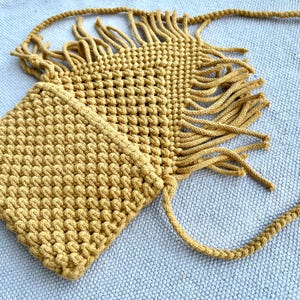 May include: A mustard yellow macrame crossbody bag featuring a textured knotted pattern, a rectangular main compartment, and long decorative fringe detailing along the edges. The bag includes a matching braided shoulder strap for carrying.