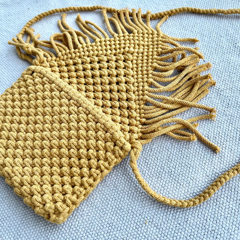 May include: A mustard yellow macrame crossbody bag featuring a textured knotted pattern, a rectangular main compartment, and long decorative fringe detailing along the edges. The bag includes a matching braided shoulder strap for carrying.