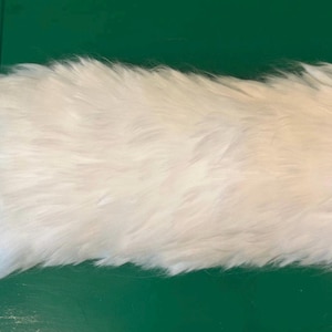 May include: A long, fluffy white faux fur tail accessory featuring a soft, dense texture and a small silver-toned metal ring attached to one end for secure fastening. The object has an elongated, tapered shape with thick, layered white synthetic fibres.