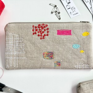 May include: A beige linen zippered pouch with colorful embroidered geometric shapes. The pouch is decorated with red, pink, yellow, blue, and white thread.