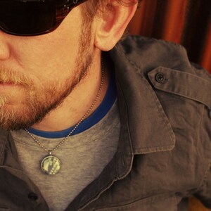 May include: A man wearing a grey t-shirt with a blue trim and a brown jacket. He is wearing a silver chain necklace with a round pendant.