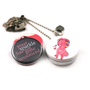 May include: A silver necklace with a charm that has a pink unicorn on one side and a black button with the text "Your sparkle has not gone unnoticed" on the other side.
