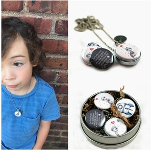 May include: A silver chain necklace with a round, white button charm featuring a black and white illustration of a small astronaut holding a red balloon. The button charm has the text "he believed he belonged among the stars and across the universe it felt like home" printed on it. The necklace is in a small, silver tin with other button charms.
