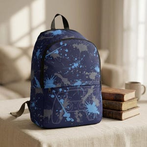 May include: Navy blue backpack with a splatter paint design in shades of blue and gray. The front of the backpack features a light blue outline of a video game controller. The backpack has a black handle and straps.