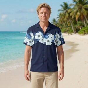 May include: Navy blue short-sleeved shirt featuring a white and blue floral pattern across the chest. The shirt has a button-down front and a collared neckline. The model is wearing khaki trousers on a beach.