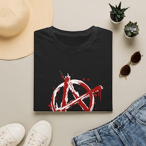 Peut inclure: T-shirt noir avec un symbole d'anarchie blanc et rouge. Le symbole est un cercle avec un "A" stylisé à l'intérieur, avec des détails de peinture rouge éclaboussée. Le t-shirt est plié sur une surface neutre.