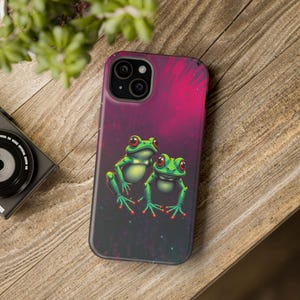 May include: A smartphone case featuring a vibrant graphic of two green frogs with large red eyes against a dark, magenta-toned background. The case has a black camera cutout area and a smooth, matte finish, showcasing a detailed illustration of amphibians.