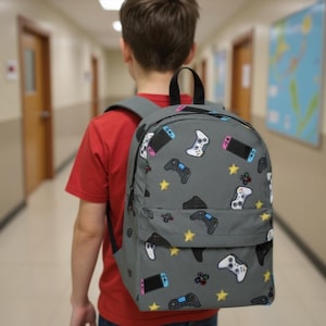 May include: A gray backpack with a pattern of video game controllers and yellow stars. The backpack has a black handle and straps. The person in the image is wearing a red shirt.