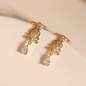 May include: A pair of gold-colored earrings with a vintage design. Each earring features an ornate, filigree-style pendant with a central clear stone and a teardrop-shaped crystal drop. The earrings are displayed on a white surface.