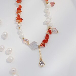 May include: A delicate necklace with a mix of red, orange, and white beads. The necklace features a large white pearl charm with a smaller teardrop-shaped crystal pendant.