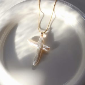 May include: A gold chain necklace with a white pearl cross pendant. The necklace is displayed on a white plate.