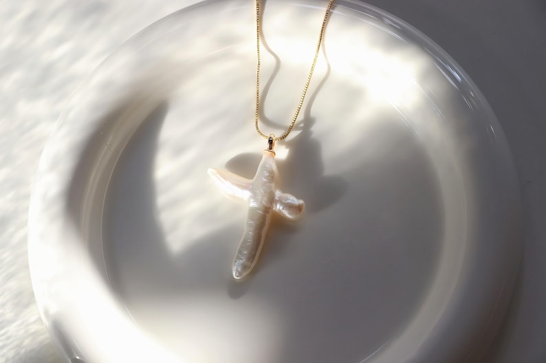 May include: A gold chain necklace with a white pearl cross pendant. The necklace is displayed on a white plate.
