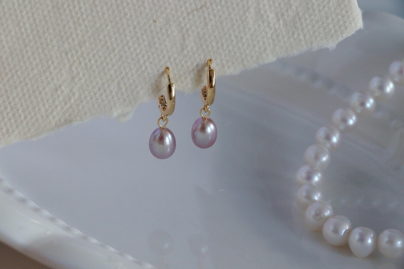 May include: A pair of gold hoop earrings with dangling, light purple, oval-shaped pearls. A white pearl necklace is partially visible in the background. The earrings are displayed on a white surface.