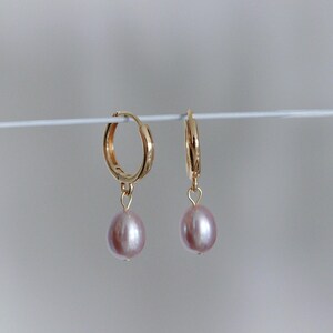 May include: A pair of gold-colored hoop earrings with a dangling, light pink, oval-shaped pearl. The earrings are displayed against a plain, light gray background. The earrings are a classic jewelry design.