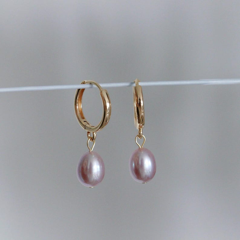 May include: A pair of gold-colored hoop earrings with a dangling, light pink, oval-shaped pearl. The earrings are displayed against a plain, light gray background. The earrings are a classic jewelry design.