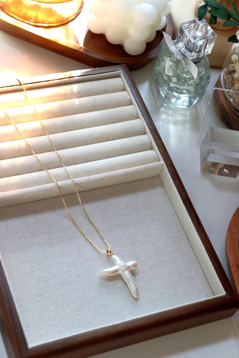 May include: A gold chain necklace with a large white pearl cross pendant. The necklace is displayed in a wooden jewellery box with a beige velvet lining.
