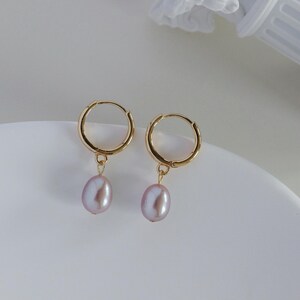 May include: A pair of gold hoop earrings with a dangling, light purple, oval-shaped pearl. The earrings are displayed on a white, curved surface. The background is a soft white.