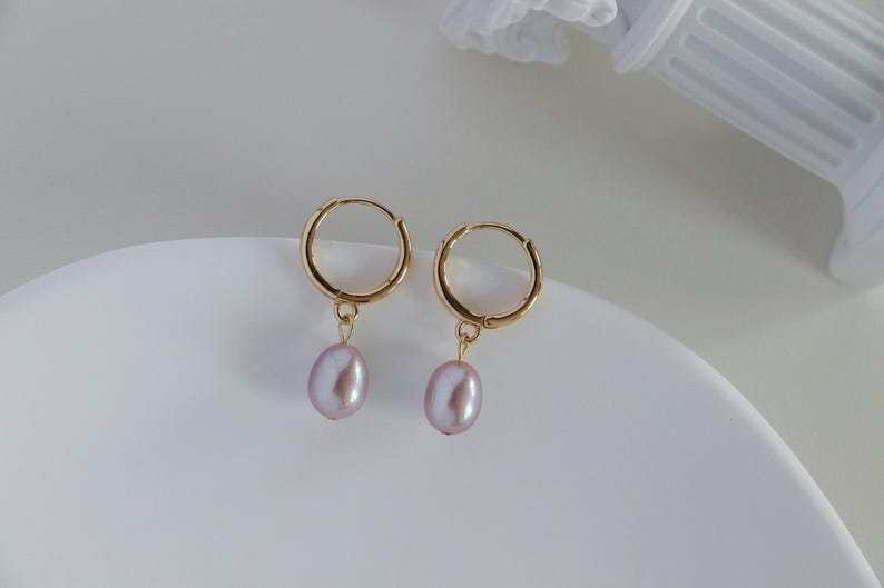 May include: A pair of gold hoop earrings with a dangling, light purple, oval-shaped pearl. The earrings are displayed on a white, curved surface. The background is a soft white.