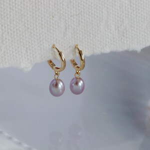 May include: A pair of gold hoop earrings with dangling lavender-colored pearls. The earrings are displayed on a white surface, with a partial view of a pearl necklace in the background. The earrings are a classic jewelry item.