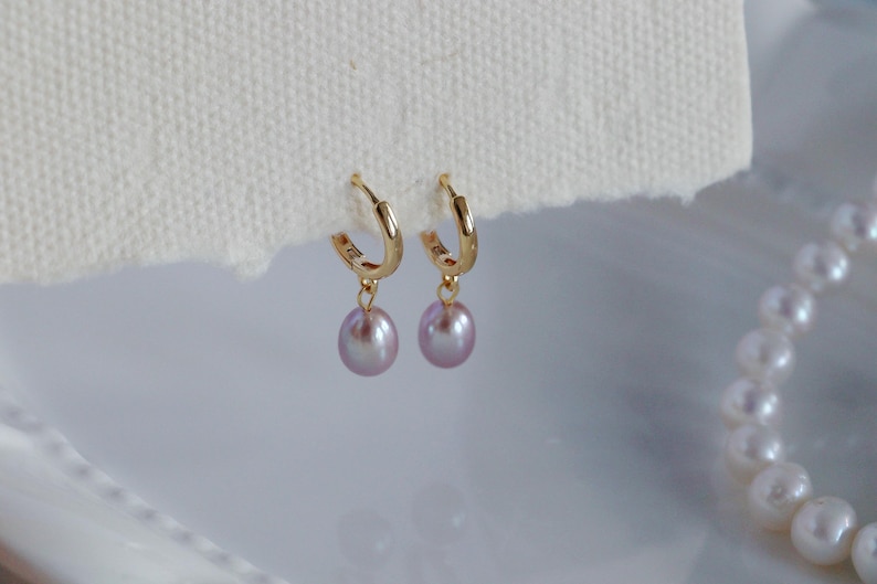 May include: A pair of gold hoop earrings with dangling lavender-colored pearls. The earrings are displayed on a white surface, with a partial view of a pearl necklace in the background. The earrings are a classic jewelry item.