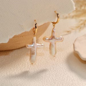 May include: A pair of gold-colored hoop earrings, each featuring a cross-shaped, off-white pearl pendant. The earrings are displayed on a neutral-toned surface with a soft, blurred background.