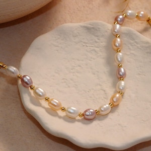 Peut inclure: Un bracelet de perles délicat avec des perles ovales blanches, roses et pêche, entrecoupées de petites perles dorées. Le bracelet est présenté sur une surface blanche texturée, avec un fermoir à chaîne dorée.