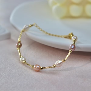Peut inclure: Un bracelet doré délicat orné d'une série de perles nacrées blanches, roses et marron. Le bracelet est présenté sur une assiette blanche, mettant en valeur son design élégant. Les perles mesurent environ 0,6 cm de diamètre.