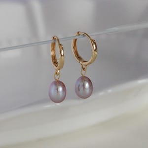 May include: A pair of gold-colored hoop earrings with a small, dangling, light purple pearl. The earrings are suspended against a soft, white background, highlighting the jewelry's delicate design and color.
