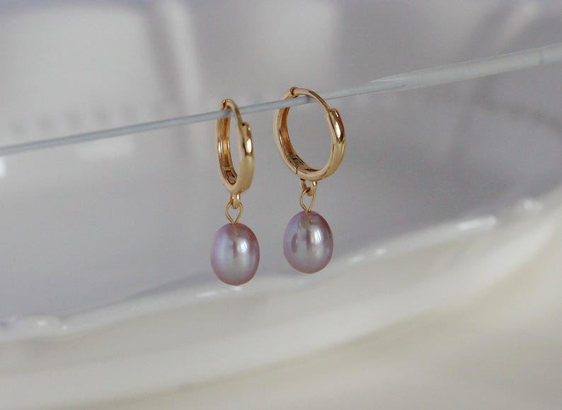 May include: A pair of gold-colored hoop earrings with a small, dangling, light purple pearl. The earrings are suspended against a soft, white background, highlighting the jewelry's delicate design and color.