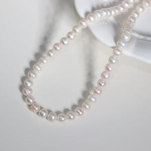 May include: A delicate pearl necklace with a soft, iridescent sheen. The necklace is composed of numerous small, round pearls, arranged in a single strand. The pearls are a creamy white color with subtle pink and lavender undertones. The necklace is displayed on a white surface.