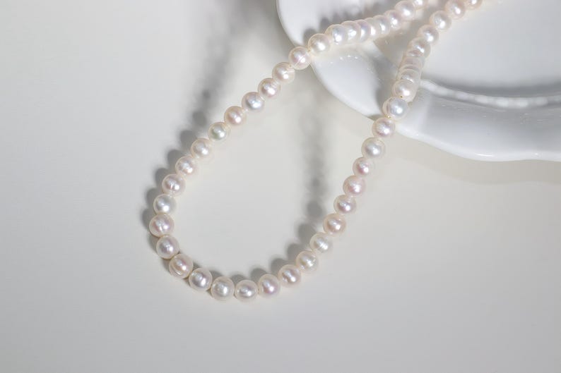 May include: A delicate pearl necklace with a soft, iridescent sheen. The necklace is composed of numerous small, round pearls, arranged in a single strand. The pearls are a creamy white color with subtle pink and lavender undertones. The necklace is displayed on a white surface.