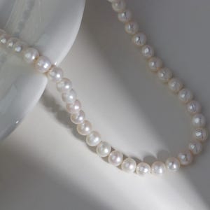 May include: A delicate pearl necklace with a classic design. The necklace features numerous round, white pearls strung together. The pearls are displayed on a white surface, creating a contrast that highlights their luster.