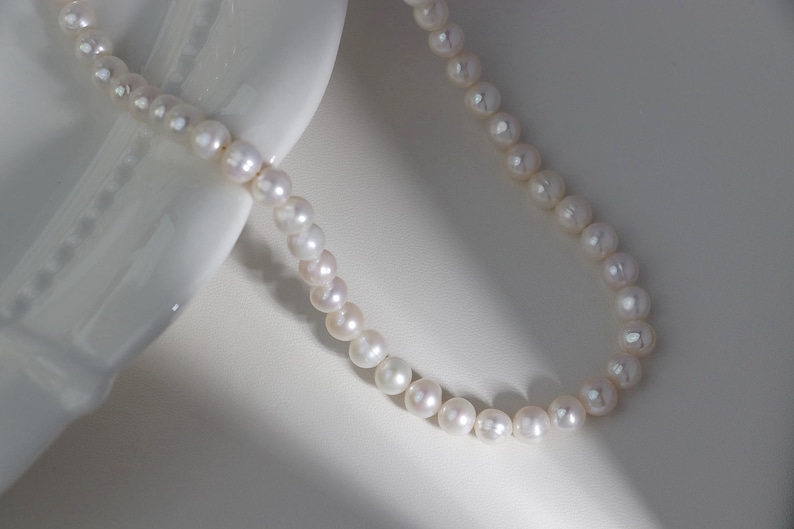 May include: A delicate pearl necklace with a classic design. The necklace features numerous round, white pearls strung together. The pearls are displayed on a white surface, creating a contrast that highlights their luster.