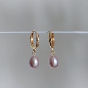 May include: A pair of gold hoop earrings with a dangling, light pink, oval-shaped pearl. The earrings are displayed against a plain, light gray background. The hoops are approximately 0.75 inches in diameter.