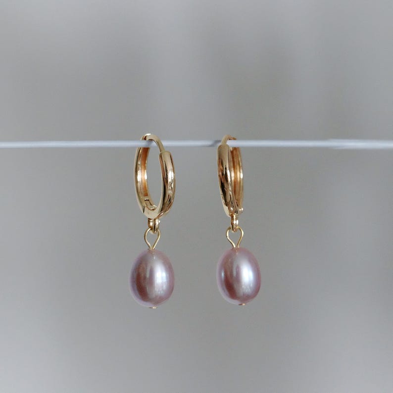 May include: A pair of gold hoop earrings with a dangling, light pink, oval-shaped pearl. The earrings are displayed against a plain, light gray background. The hoops are approximately 0.75 inches in diameter.