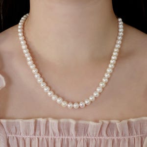 May include: A classic pearl necklace composed of numerous round, white pearls. The necklace is draped around a person's neck, against a soft pink background. The pearls are evenly sized and closely strung together.