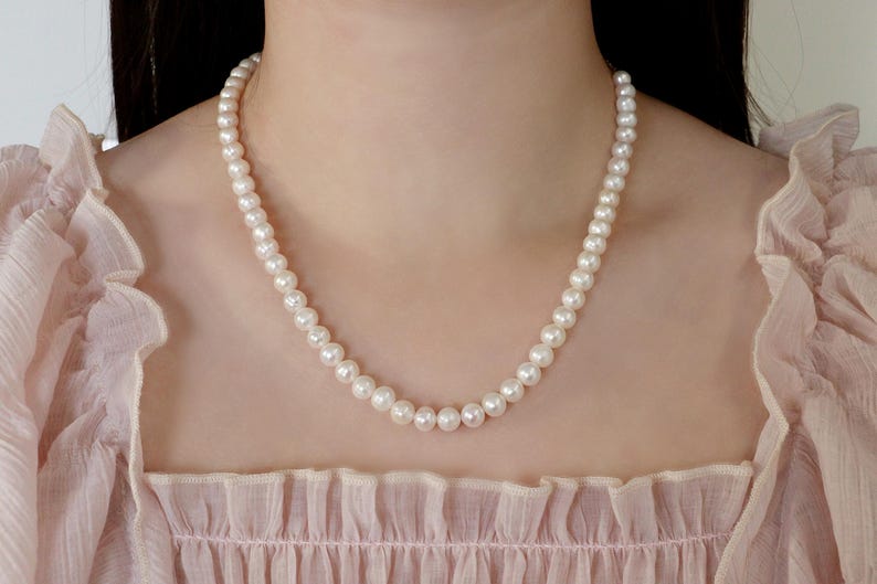 May include: A classic pearl necklace composed of numerous round, white pearls. The necklace is draped around a person's neck, against a soft pink background. The pearls are evenly sized and closely strung together.
