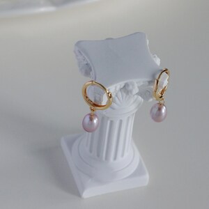 May include: A pair of gold hoop earrings with a light pink, teardrop-shaped pearl pendant. The earrings are displayed on a white, decorative column. The column has a square base and a fluted design.