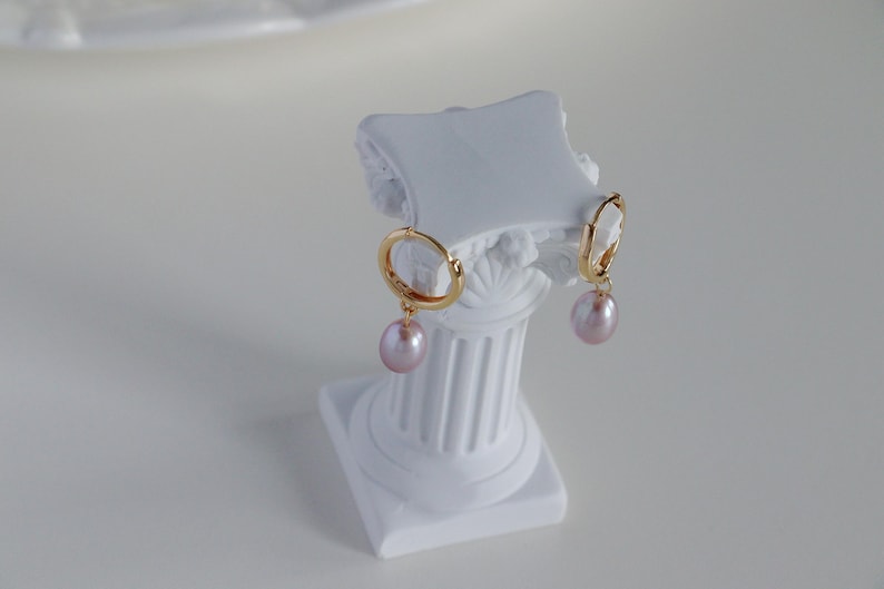 May include: A pair of gold hoop earrings with a light pink, teardrop-shaped pearl pendant. The earrings are displayed on a white, decorative column. The column has a square base and a fluted design.