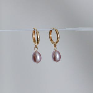 May include: A pair of gold-colored hoop earrings, each with a dangling, oval-shaped, light purple pearl. The earrings are displayed against a plain, light background, highlighting the jewelry's design and color.