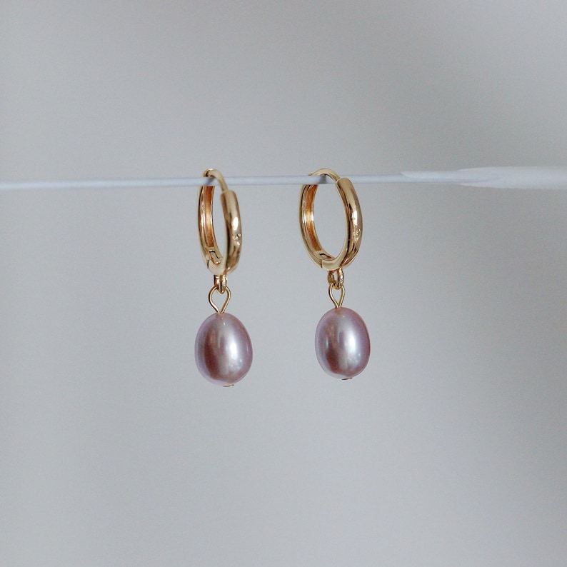May include: A pair of gold-colored hoop earrings, each with a dangling, oval-shaped, light purple pearl. The earrings are displayed against a plain, light background, highlighting the jewelry's design and color.