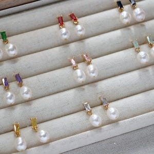 May include: An assortment of pearl earrings with coloured gemstone details. The earrings showcase white pearls combined with gold-coloured metal and rectangular gemstones in green, red, purple, pink, and yellow. The earrings are displayed on a light-coloured fabric.