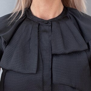 May include: A black, textured top with a unique collar design. The top features a round neckline with a single button closure and a layered, pleated detail around the shoulders. The fabric has a subtle, woven pattern.
