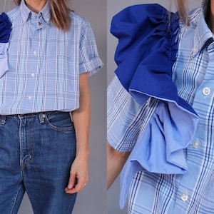 May include: A blue and white plaid short-sleeve shirt with a unique design. The shirt features a layered, ruffled accent on one shoulder, with shades of blue. The model is wearing blue jeans.