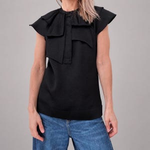 May include: A black textured top with a unique ruffled collar and short sleeves. The top has a distinctive design with layered fabric details around the neckline and a buttoned closure. The garment is paired with blue jeans.