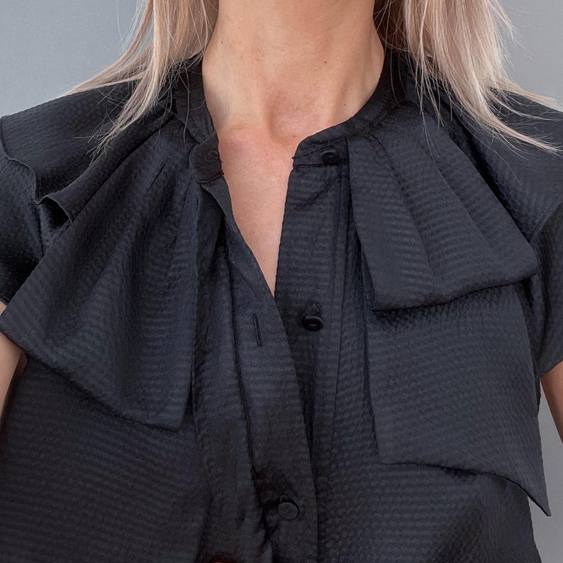 May include: A black short-sleeved blouse with a textured pattern. The blouse features a high collar and a large, layered bow detail at the neckline. The buttons are black and the fabric has a subtle sheen.