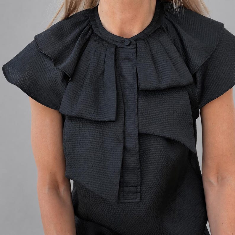 May include: Black textured short-sleeve top with a round neckline and a button closure. The top features layered ruffles around the shoulders and a pleated bib detail down the front. The fabric has a subtle, grid-like pattern.